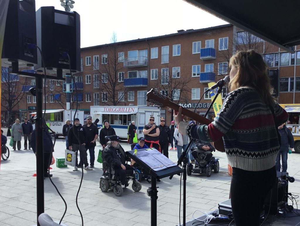 En ung, kvinnlig trubadur med långt ljust hår spelar på gitarr och sjunger inför publik. på torget i Skellefteå