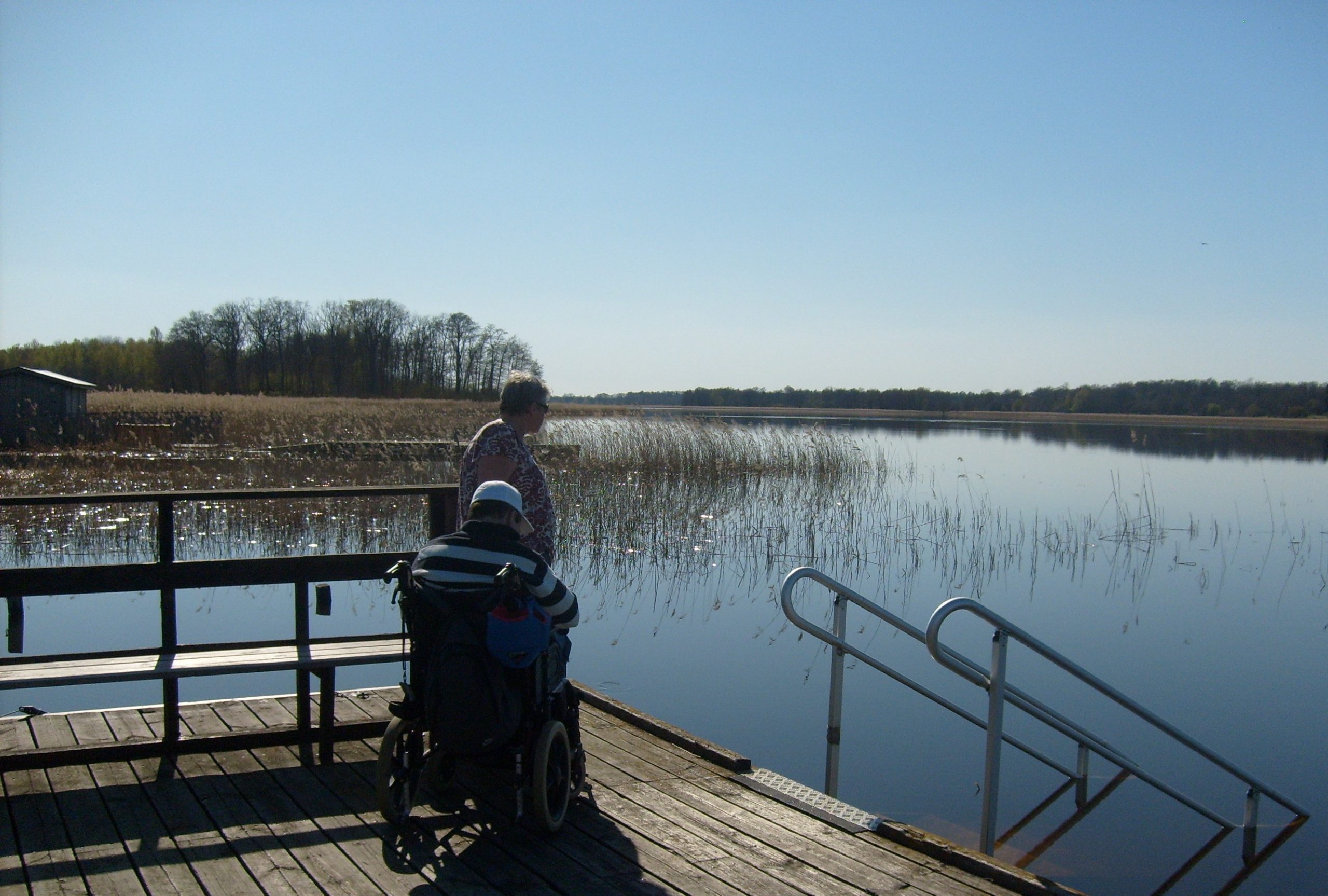 Kvinna och en man i rullstol på en brygga med ramp till sjön.