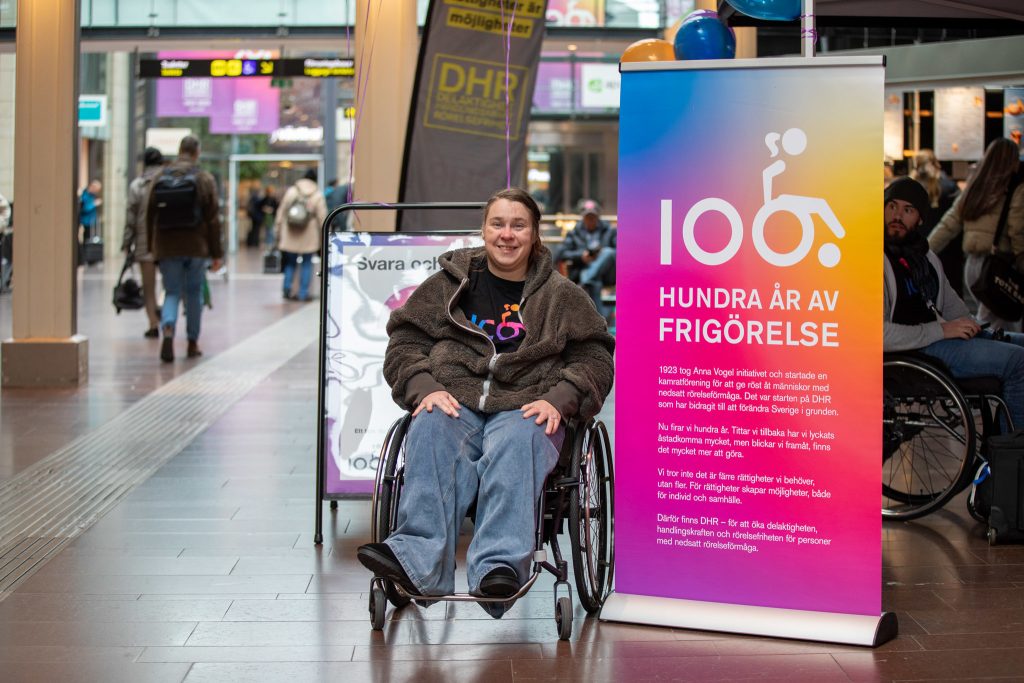 På Göteborgs Centralstation sitter en kvinna i rullstol intill en roll-up med texten "DHR 100 år av frigörelse."