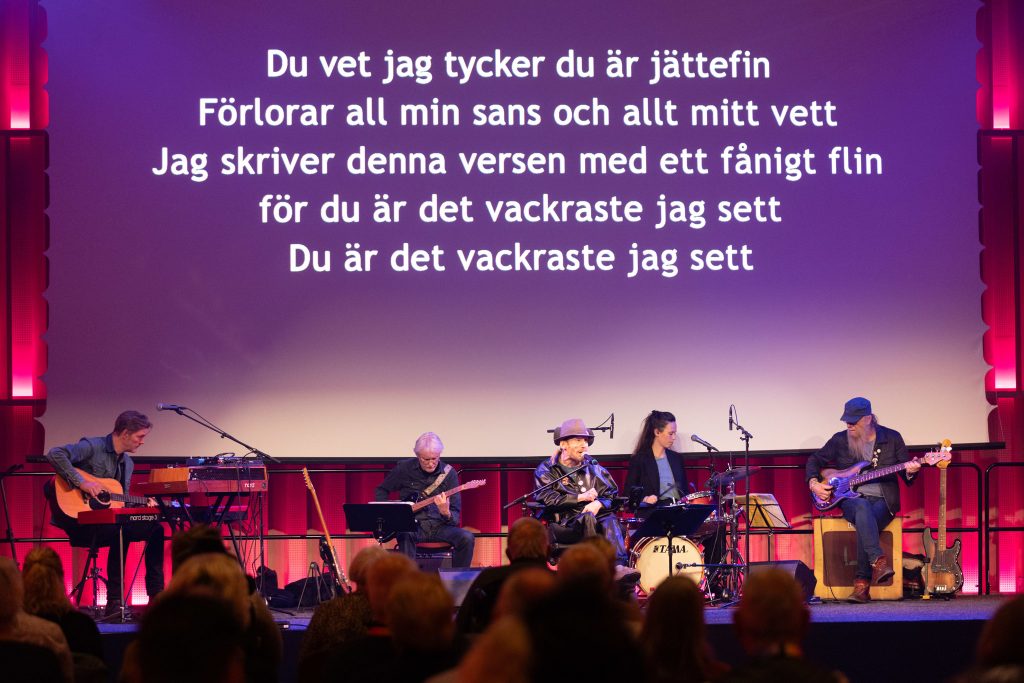 Under en text projicerad på väggen sitter Jesper Odelberg tillsammans med sina musiker.