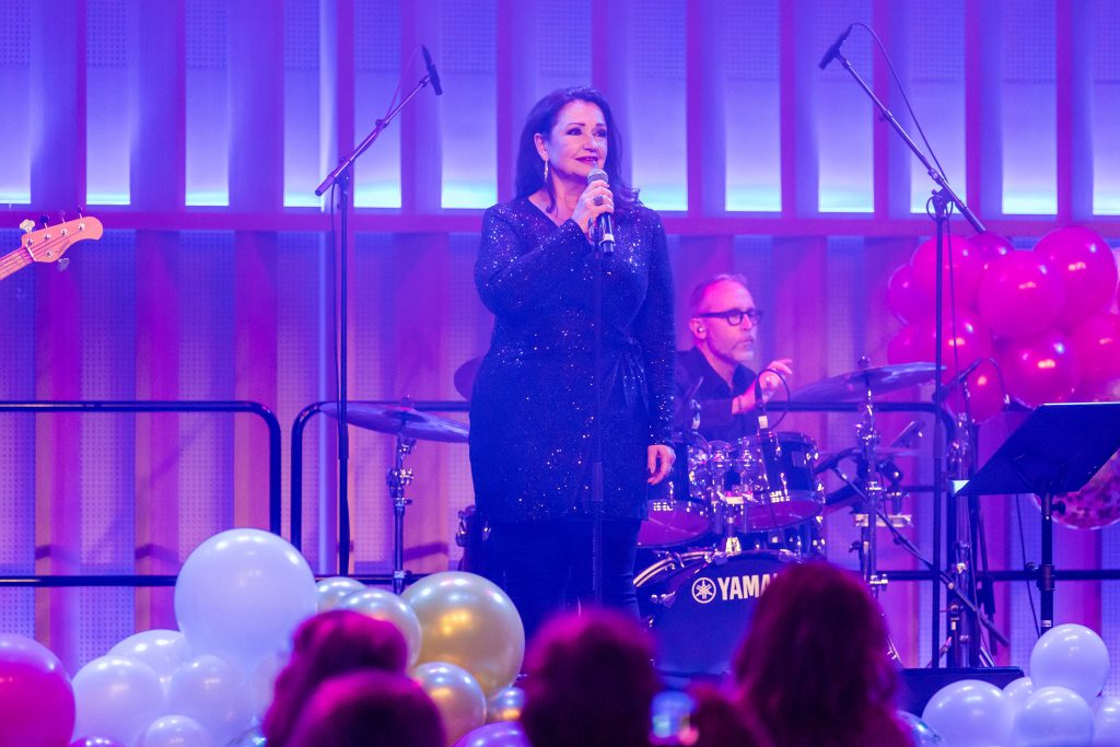 Bakom en driva av färgglada ballonger står en festklädd kvinna - Maria Möller - och sjunger i en mikrofon.
