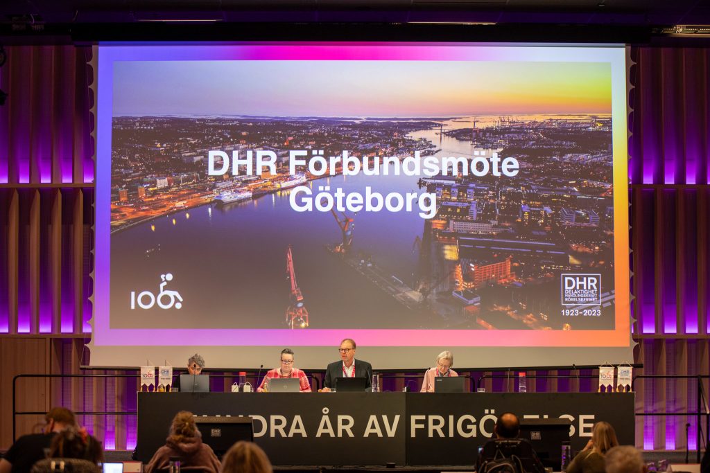 Vid ett podie under en bild av Göteborg med texten "DHR Förbundsmöte Göteborg" sitter ett presidium bestående av en man och fyra kvinnor. 