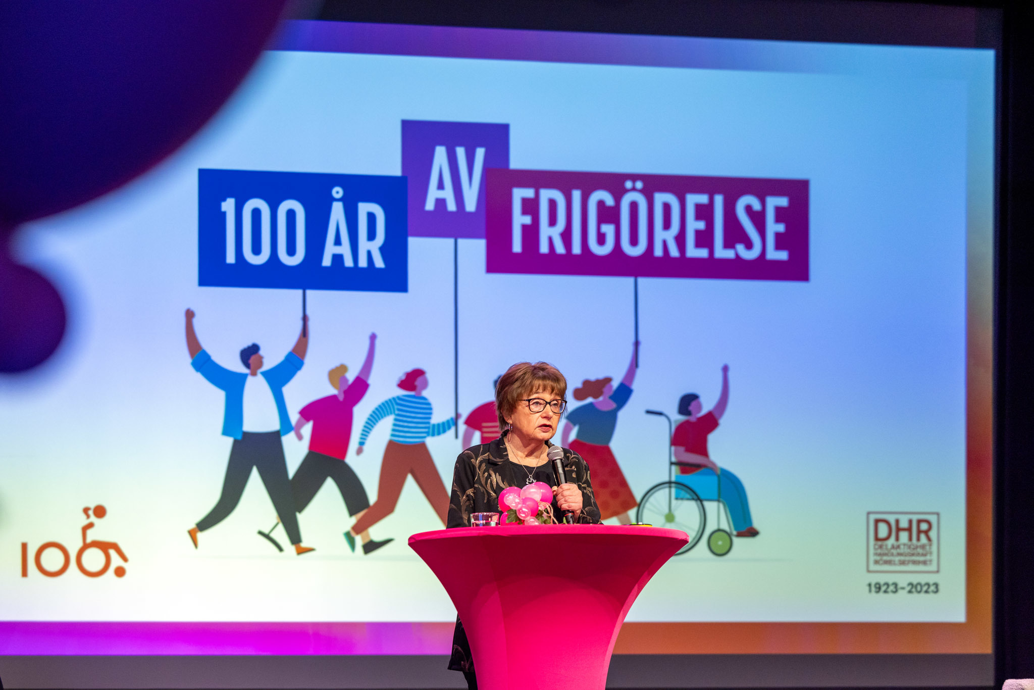 Framför texten 100 år av frigörelse" står en kvinna i mörkt, kortklippt hår och glasögon vid ett talarpodium.