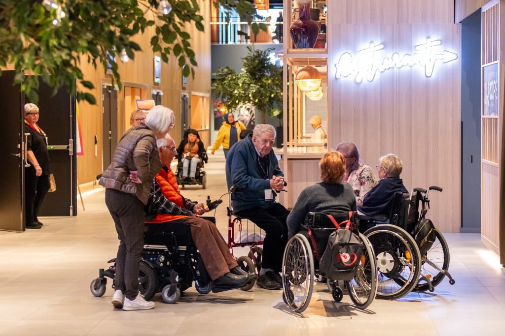 I en hotellfoajé pågår ett samtal mellan en man som sitter på sin rullator och personer som sitter i rullstol, samt stående personer. 