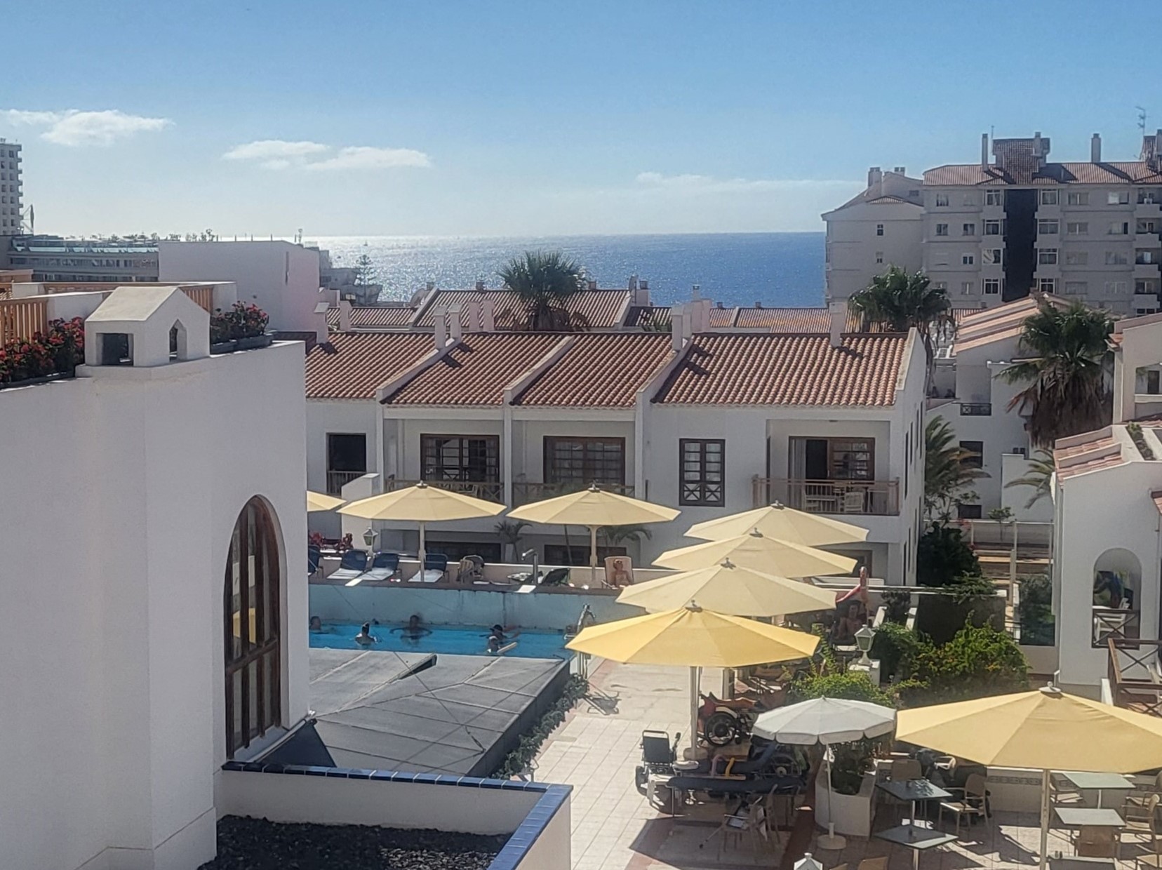 Vitrappade, lägre hus med röda tegeltak omger en pool med badande hotellgäster. Bakom husen skymtar havet.